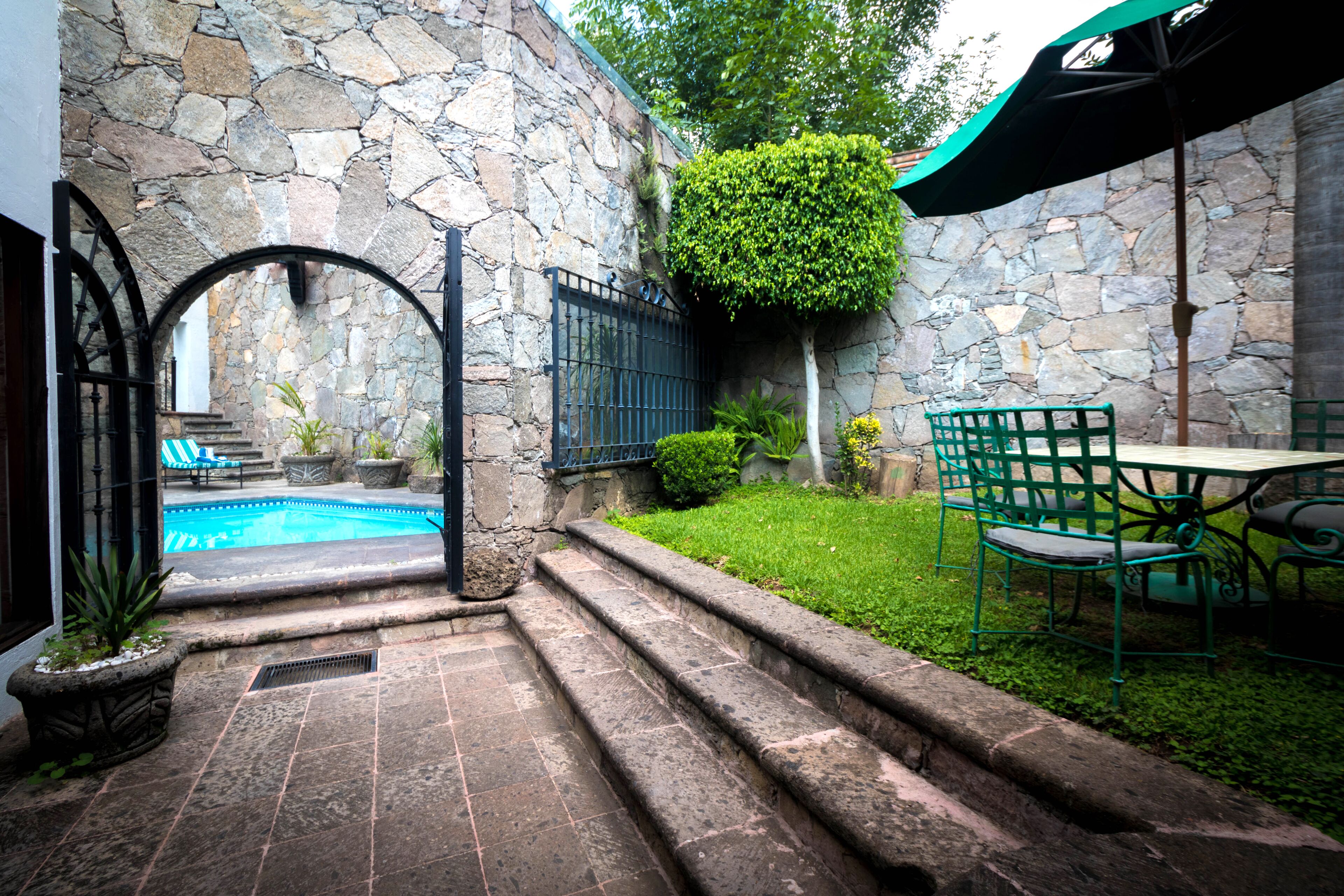 Un patio con paredes de piedra, una piscina, escalones que conducen a una zona de estar y un paraguas verde.