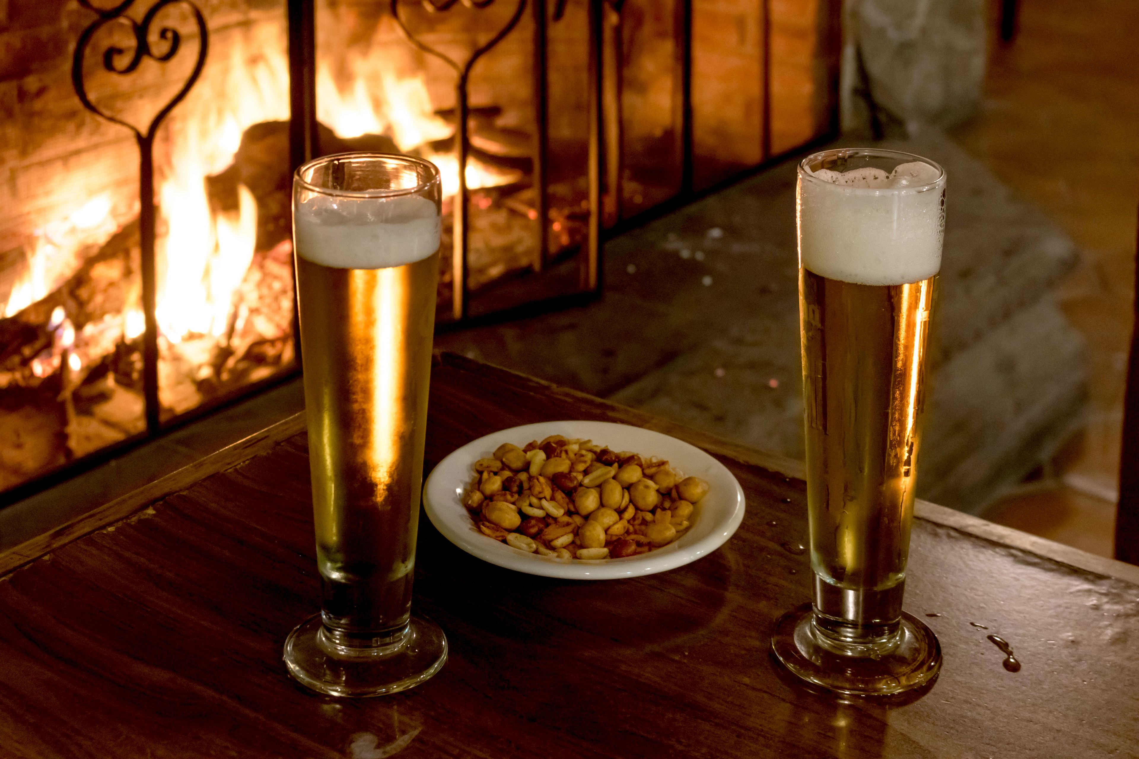 Dues copes altes de cervesa sobre una taula de fusta, amb un plat de fruits secs a prop, davant de la llar de foc.