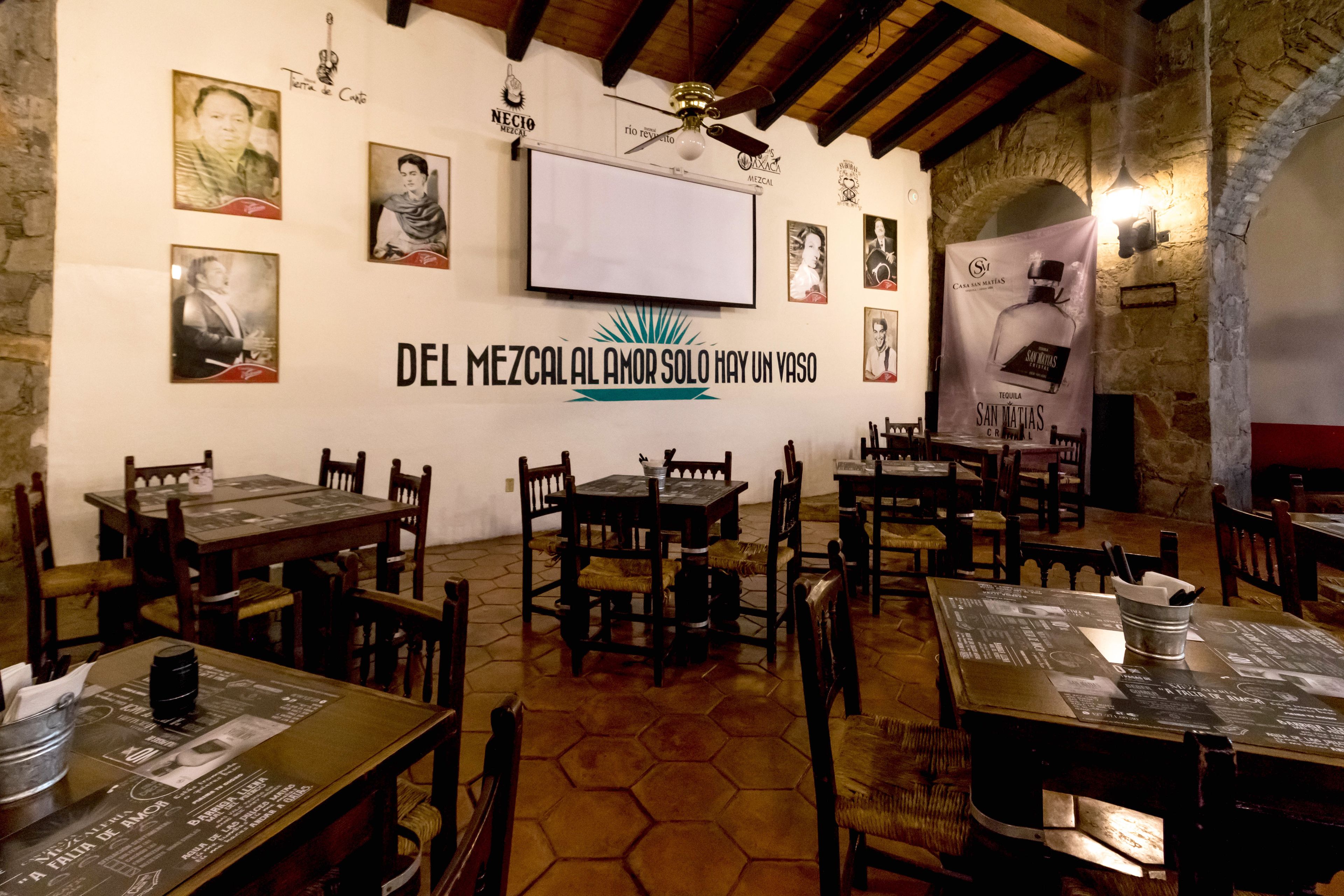 Interior de un restaurante con mesas y sillas de madera, una pantalla grande y varios carteles en las paredes.