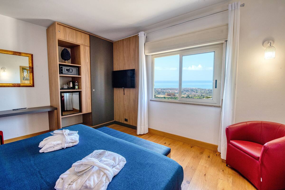 A hotel room with two beds, a red chair, a TV, a wooden cabinet, and a large window with a city view.