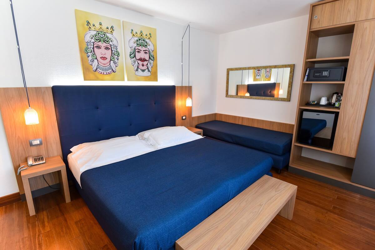 A hotel room with a large bed, bedside tables, a bench, a wooden cabinet, and artwork on the walls.