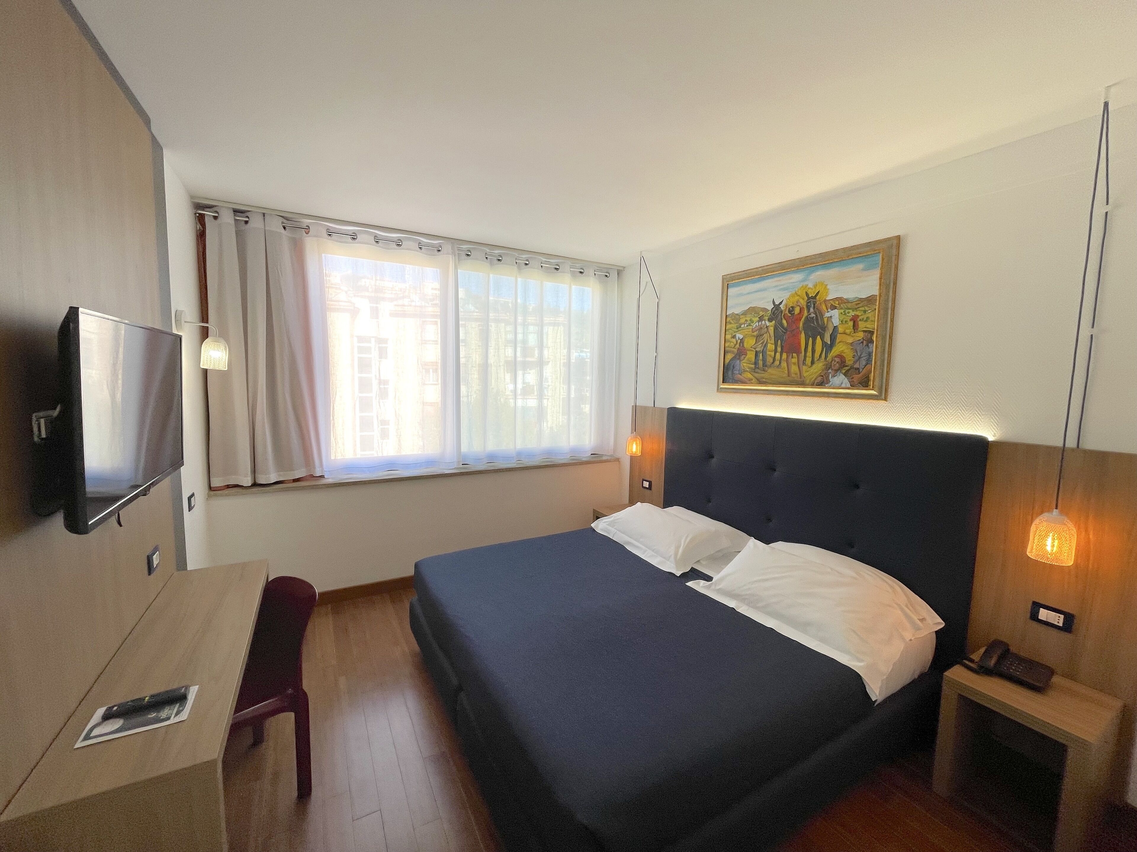 A hotel room with a bed, a desk, a television, a painting, and a window with curtains.