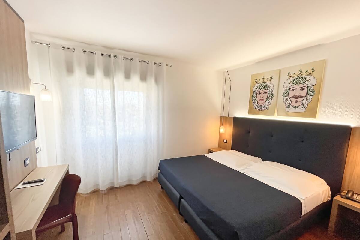 A hotel room with a bed, a desk, a chair, a TV, and artwork on the wall.