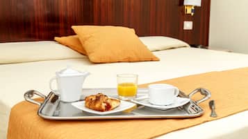 Un vassoio per la colazione ben apparecchiato, con croissant, caffè e succo d'arancia, su un letto con una testiera in legno e un solo cuscino.