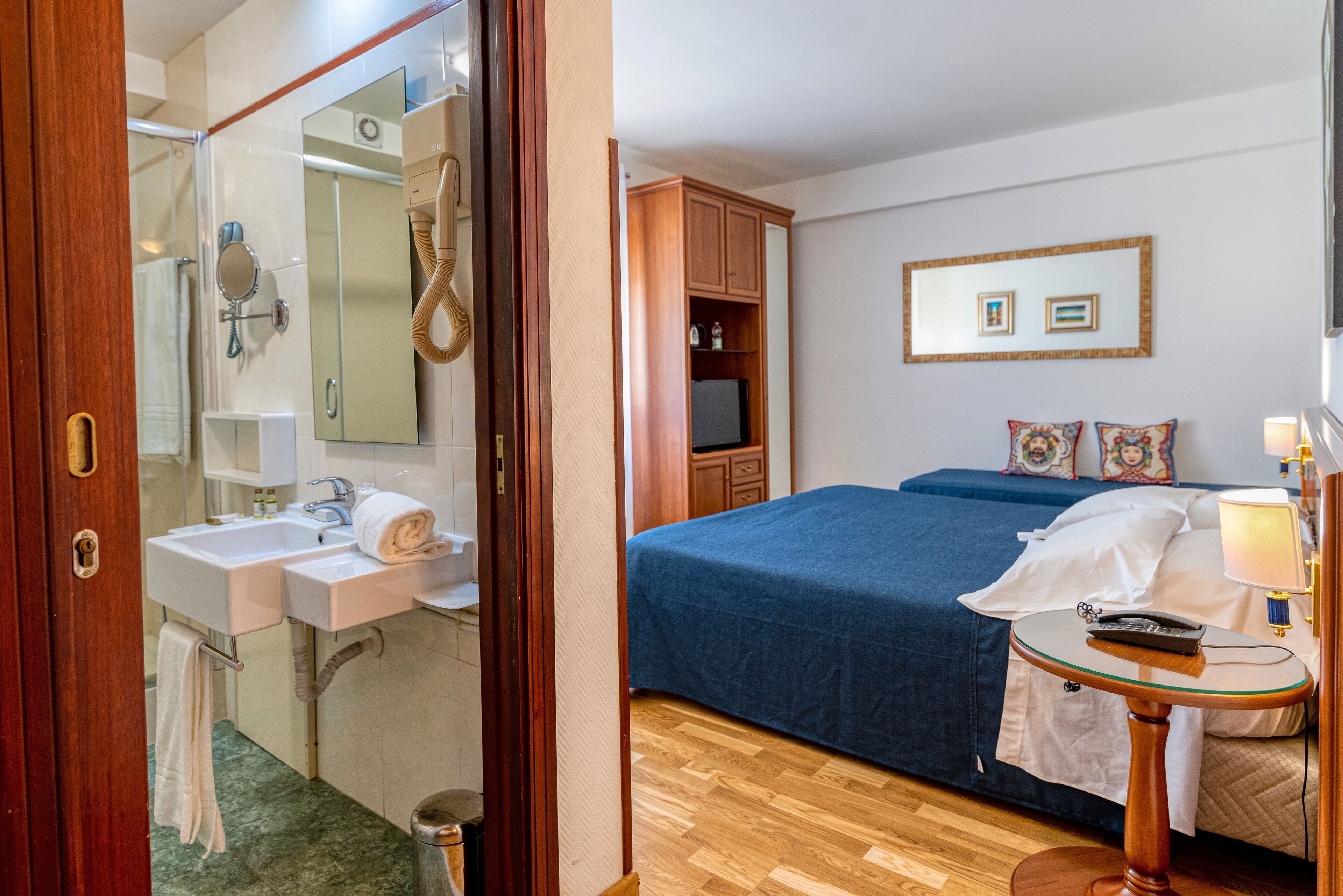Habitación de hotel con suelo de madera, una cama con colcha azul, mesita de noche con lámpara, televisor y armario de madera.