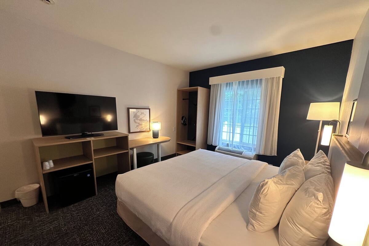 A hotel room with a large bed, a flat-screen TV, a desk with a chair, a window with curtains, and a wall-mounted lamp.