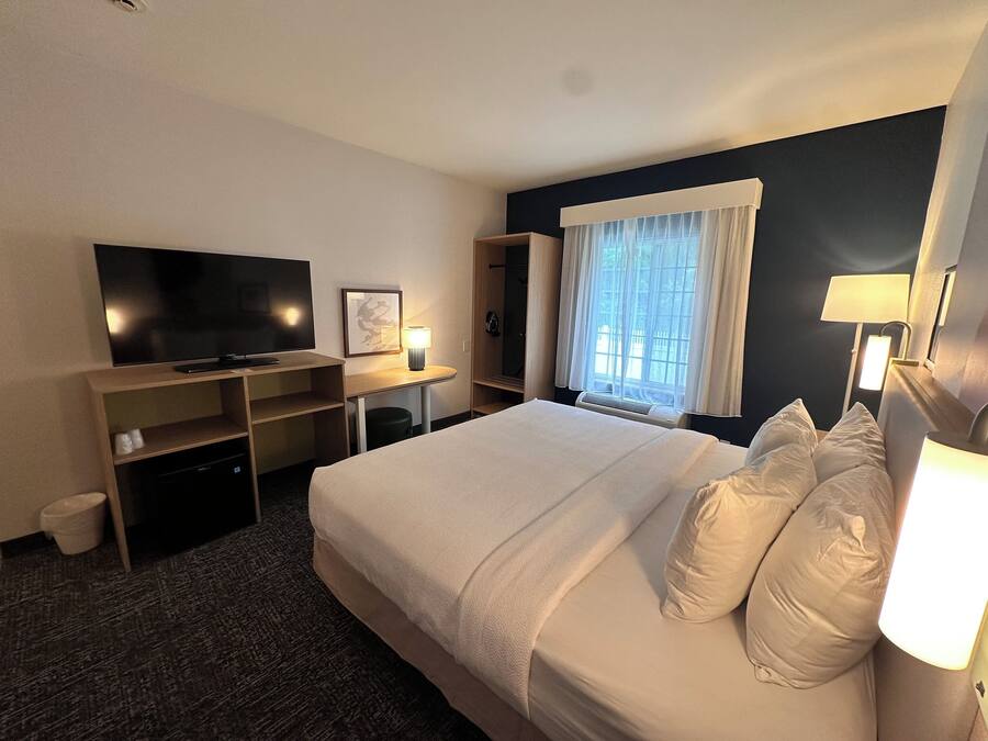 A hotel room with a large bed, a flat-screen TV, a desk with a chair, a window with curtains, and a wall-mounted lamp.