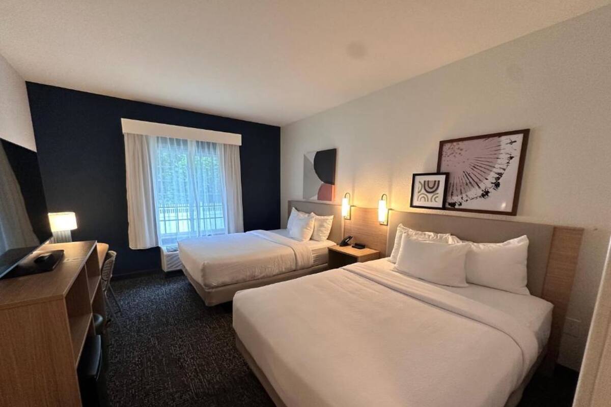 A hotel room with two beds, a desk, a chair, a window with curtains, wall-mounted lights, and a framed picture.