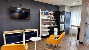 A modern living room with a flat-screen TV, a small table, a yellow armchair, a rocking chair, and a mini-fridge stocked with snacks. - Spark by Hilton Guilford (Guilford)