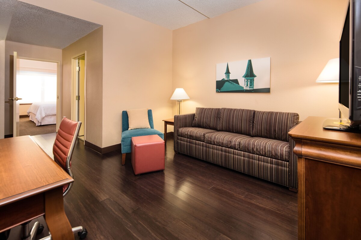 A hotel room with a wooden dining table, a striped sofa, a pink ottoman, a teal chair, and a lamp.
