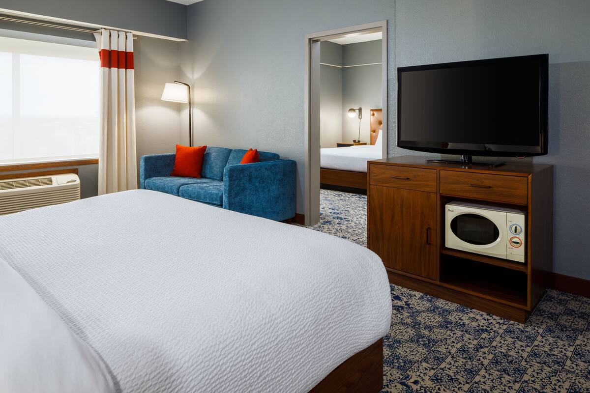 A hotel room with a bed, a blue sofa with red cushions, a television, a wooden cabinet with a microwave, and a large mirror.