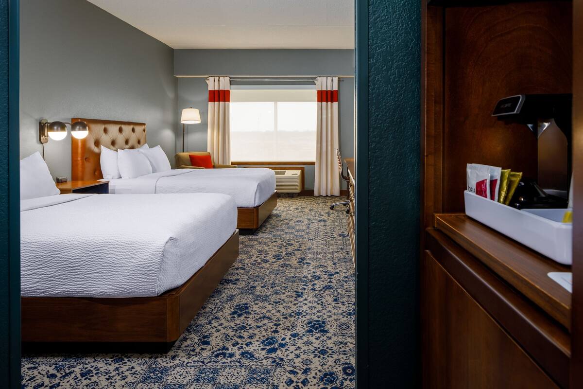 A hotel room with two beds, a wooden headboard, a bedside table with a lamp, a window with curtains, and a small seating area.