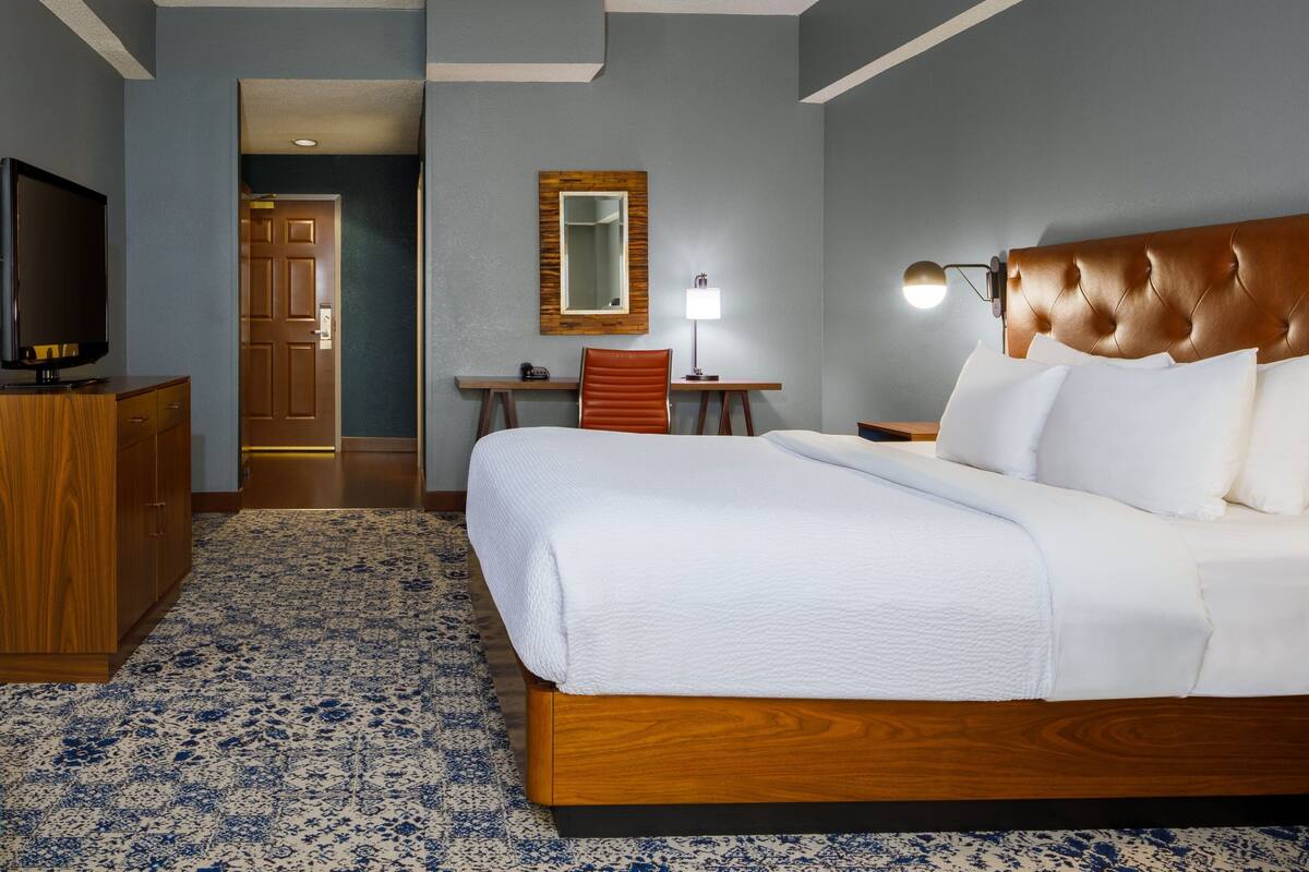 A hotel room with a large bed, a wooden headboard, a desk with a chair, a television, and a mirror on the wall.