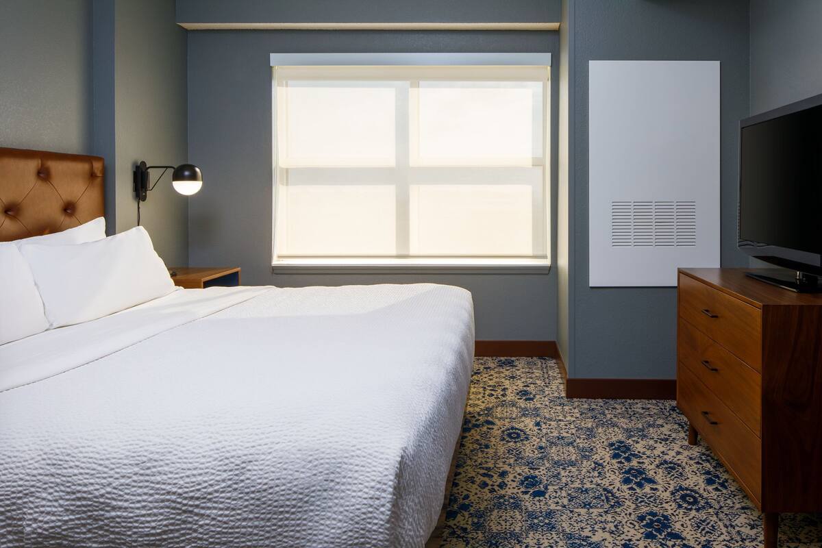 A hotel room with a large bed, a wooden nightstand, a desk with a television, a window with blinds, and a wall-mounted air conditioning unit.