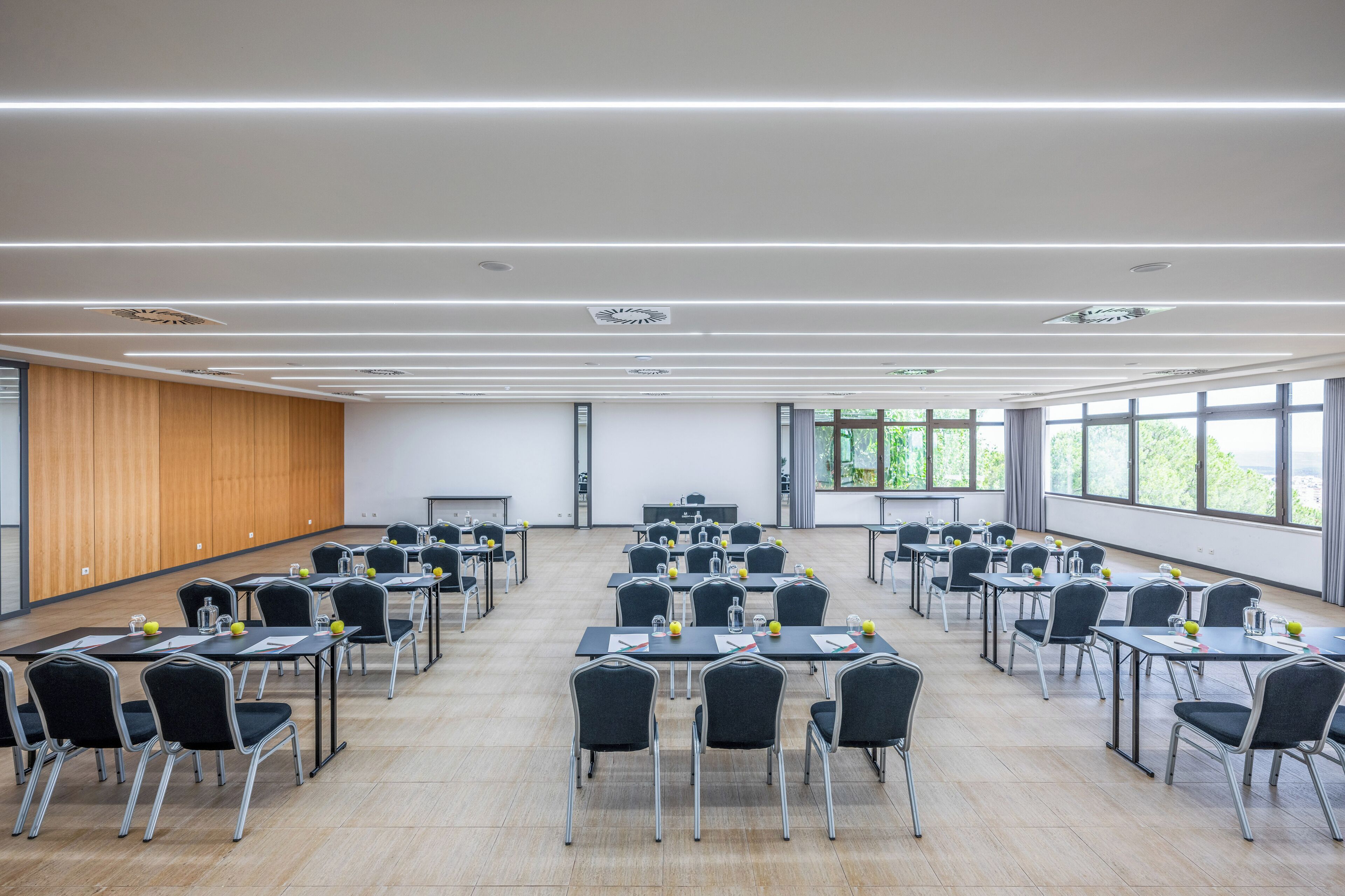 Un salón de conferencias con varias mesas y sillas, ventanas amplias y un panel de pared de madera.