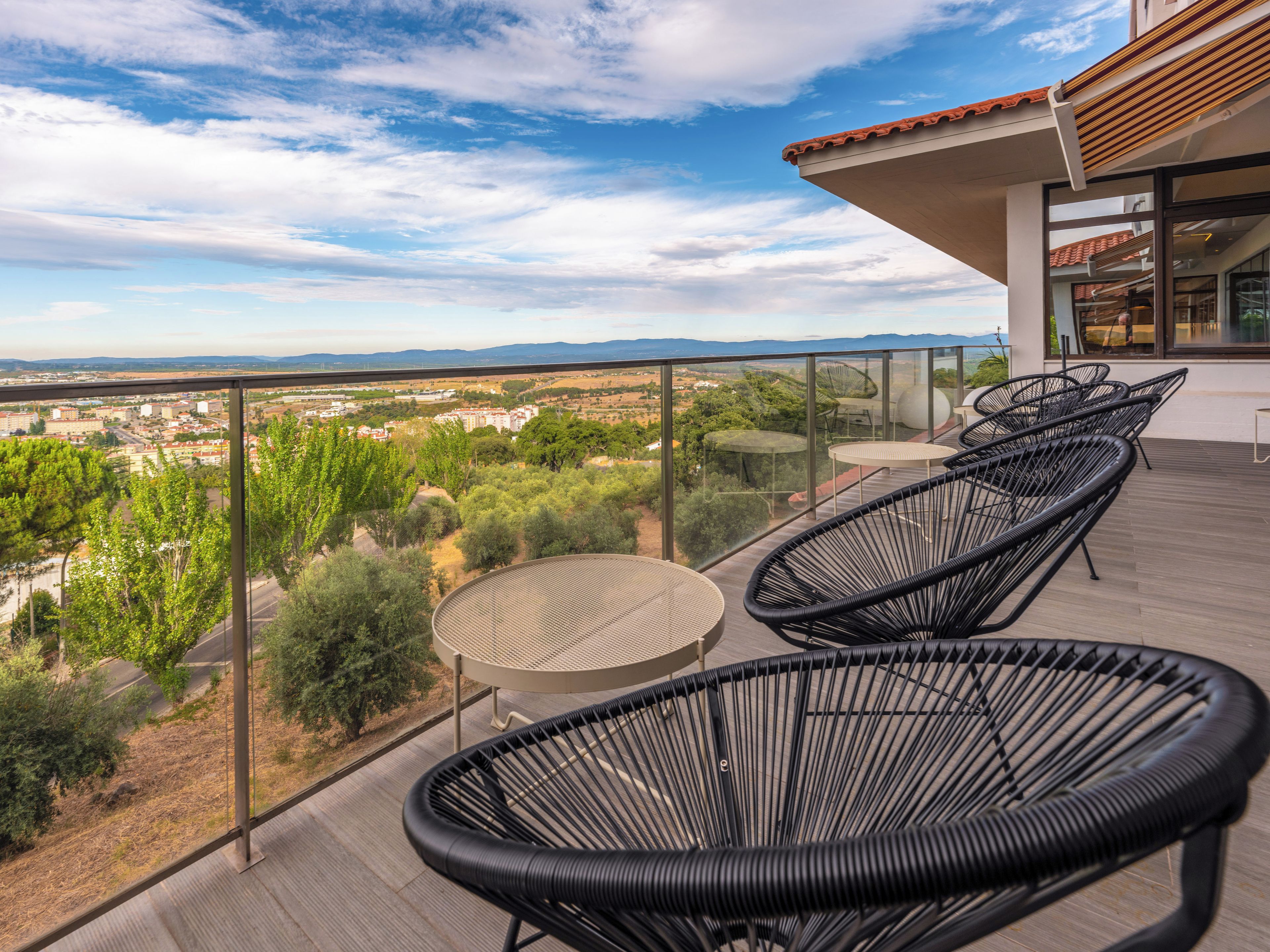 A balcony with a glass railing, wicker chairs, a round table, and a view of a cityscape and mountains.