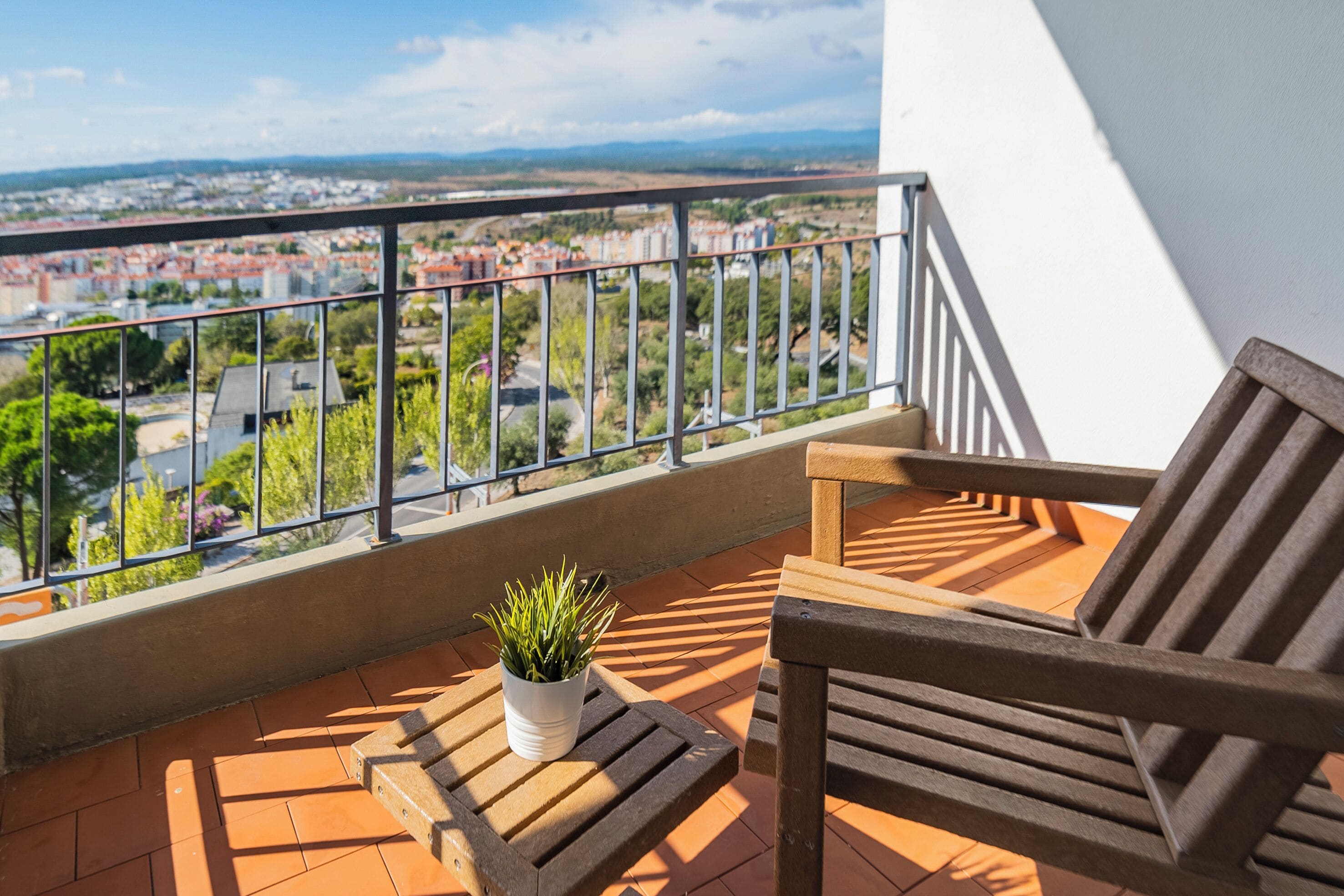 Un balcón con vistas a un paisaje urbano, una silla de madera, una planta pequeña en maceta y un suelo de baldosas.