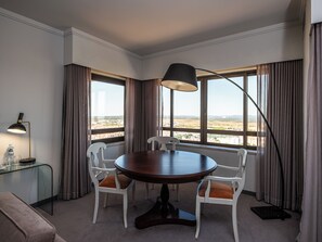 Un coin repas avec une table ronde, des chaises blanches, une table d’appoint en verre avec une lampe, et une vue sur le paysage grâce à de grandes fenêtres.