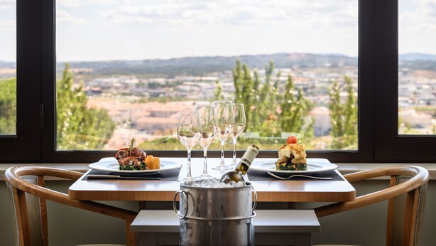A dining table with a view of a cityscape through large windows, a bottle of champagne, and two plates of food. - Melia Castelo Branco (Castelo Branco)