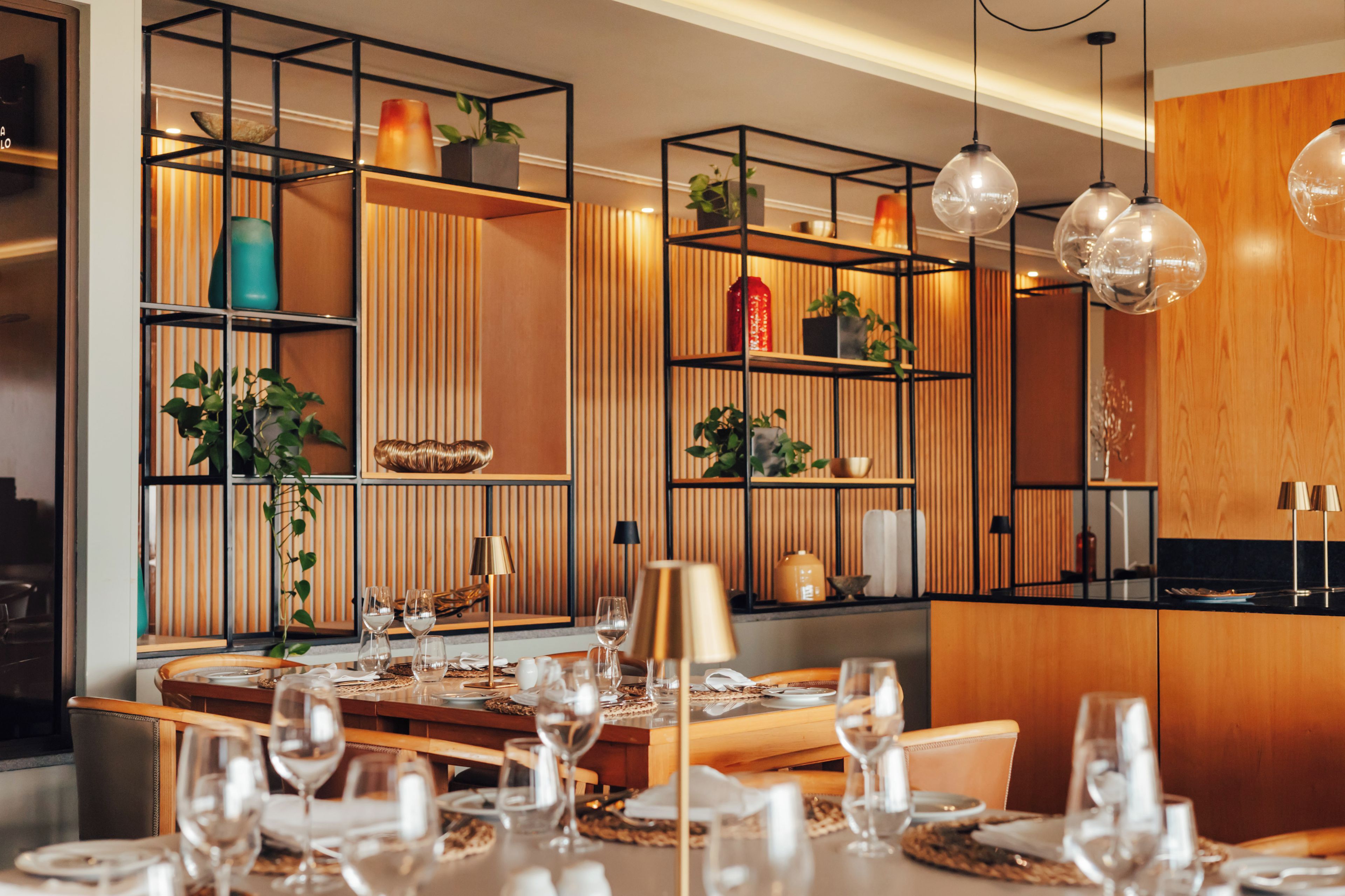 Un comedor moderno con una mesa de madera dispuesta para una comida, rodeado de estantes con plantas y objetos decorativos.