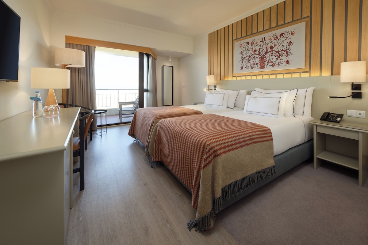 A hotel room with a large bed, a desk, and a chair. There is a window with curtains, a painting on the wall, and a lamp on the desk.