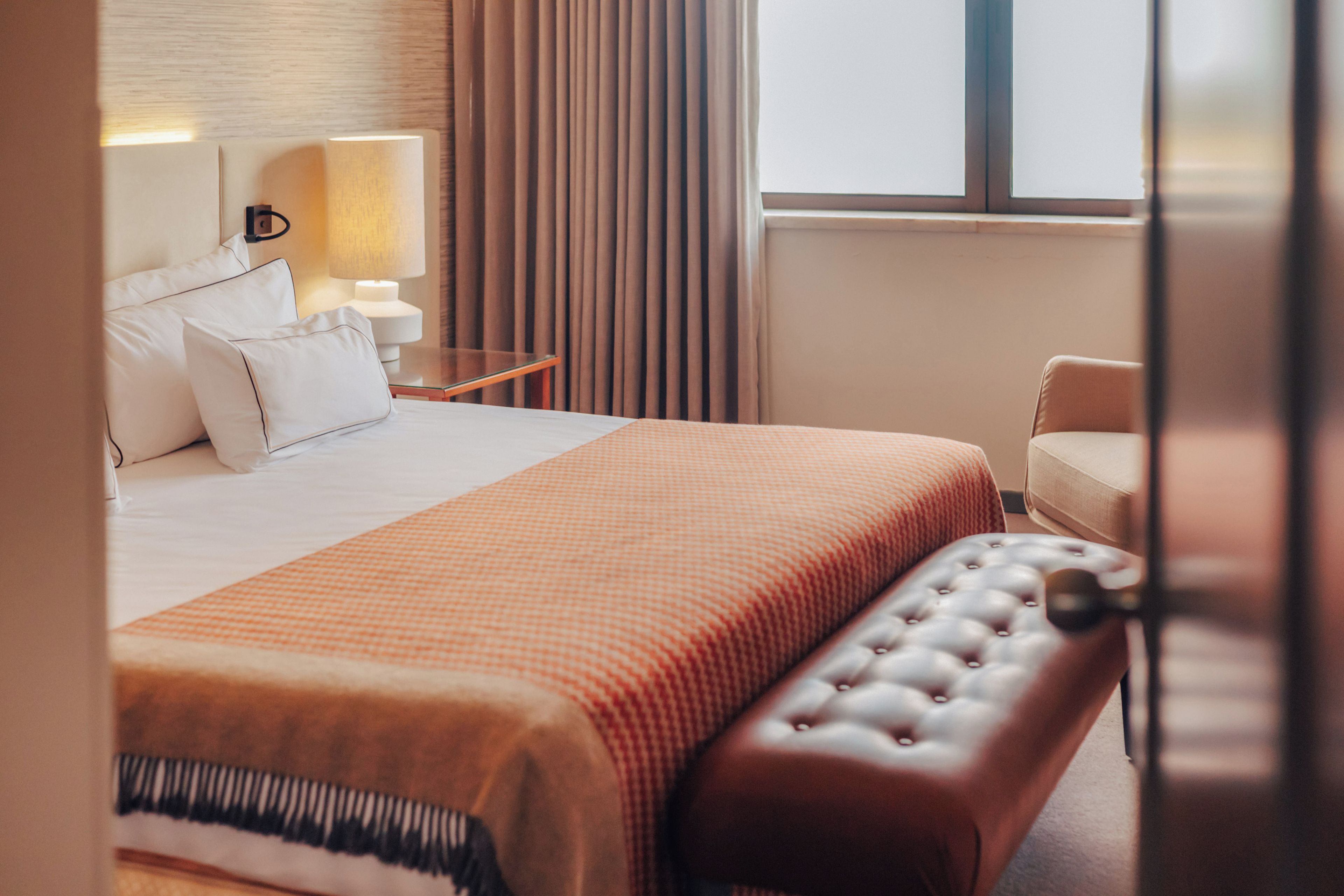 A hotel room with a bed, a nightstand, a lamp, a chair, and a window with curtains.