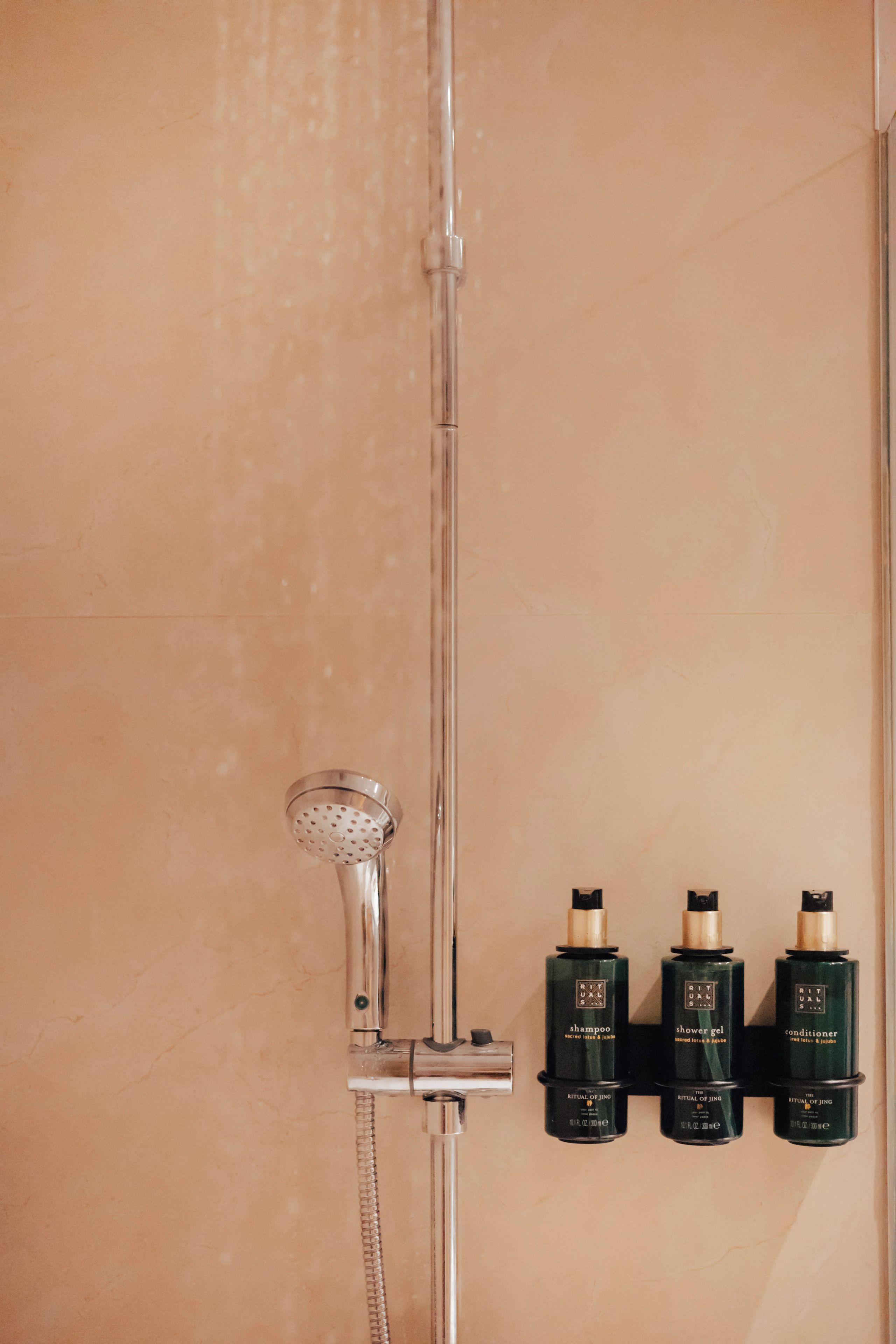 Una ducha con cabezal de ducha de mano y grifo empotrado. Hay tres botellas de champú, gel de ducha y acondicionador en una repisa.