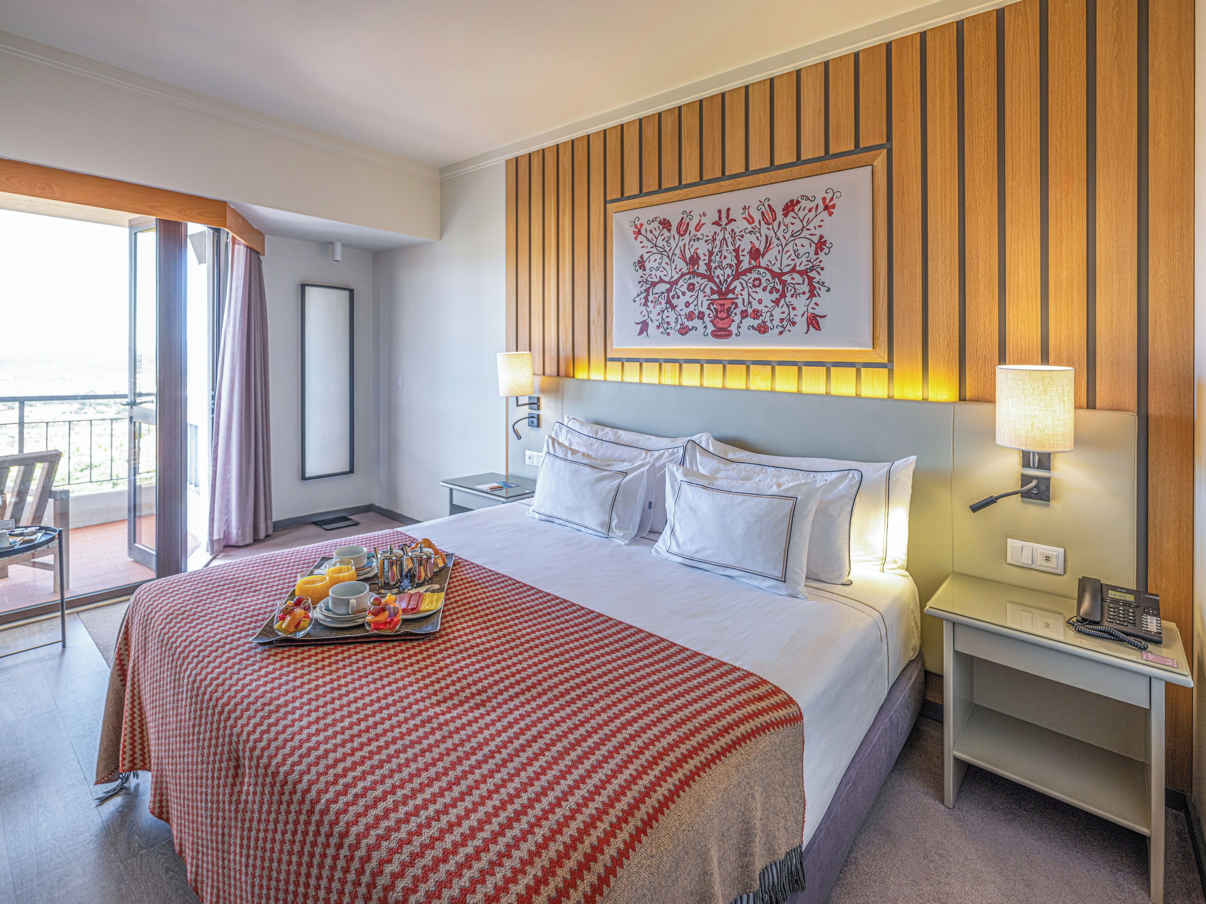 Una habitación de hotel con cama, bandeja de desayuno, mesita de noche, lámpara y vistas al exterior.