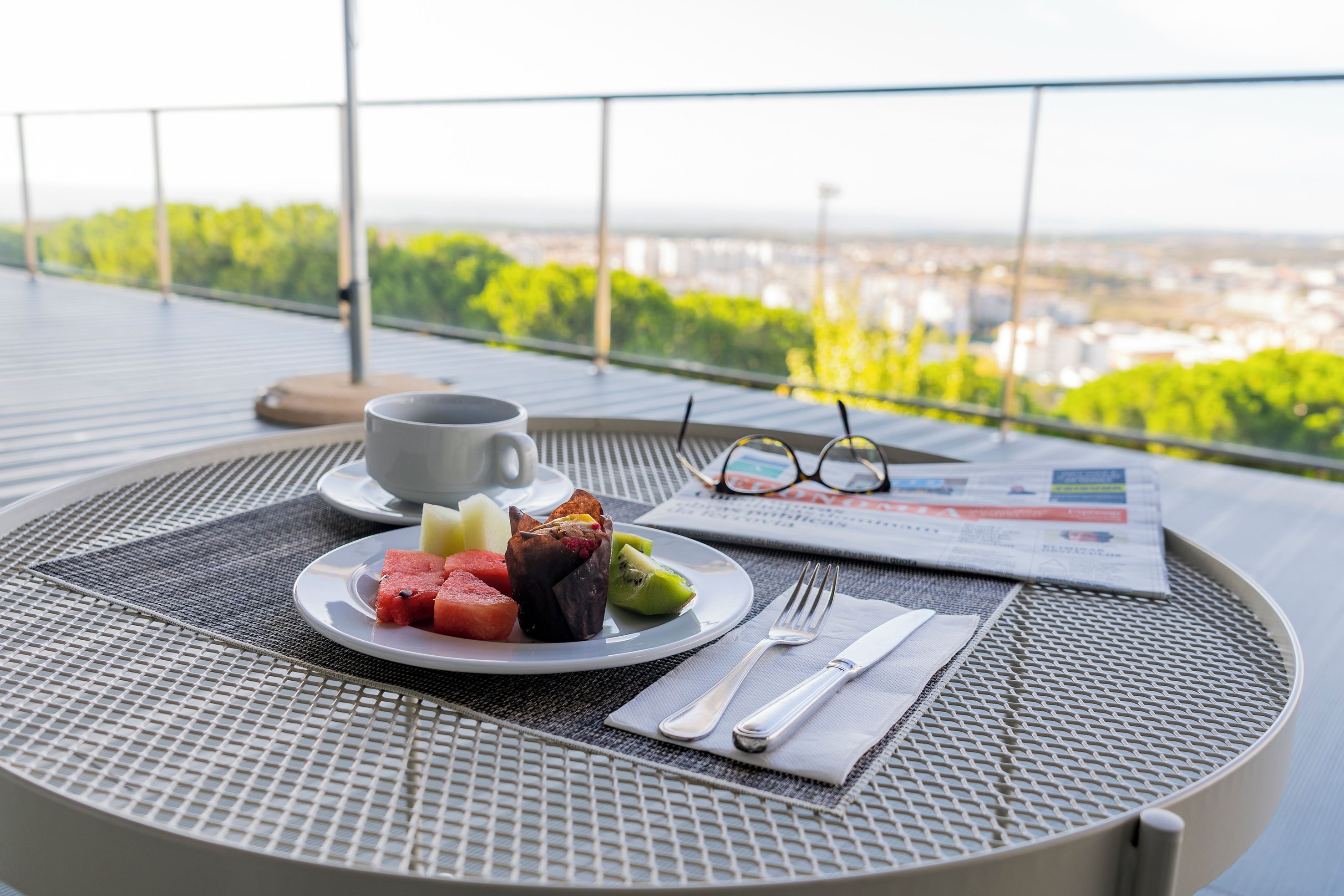A table with a plate of food, a cup, glasses, and cutlery on a balcony overlooking a cityscape.