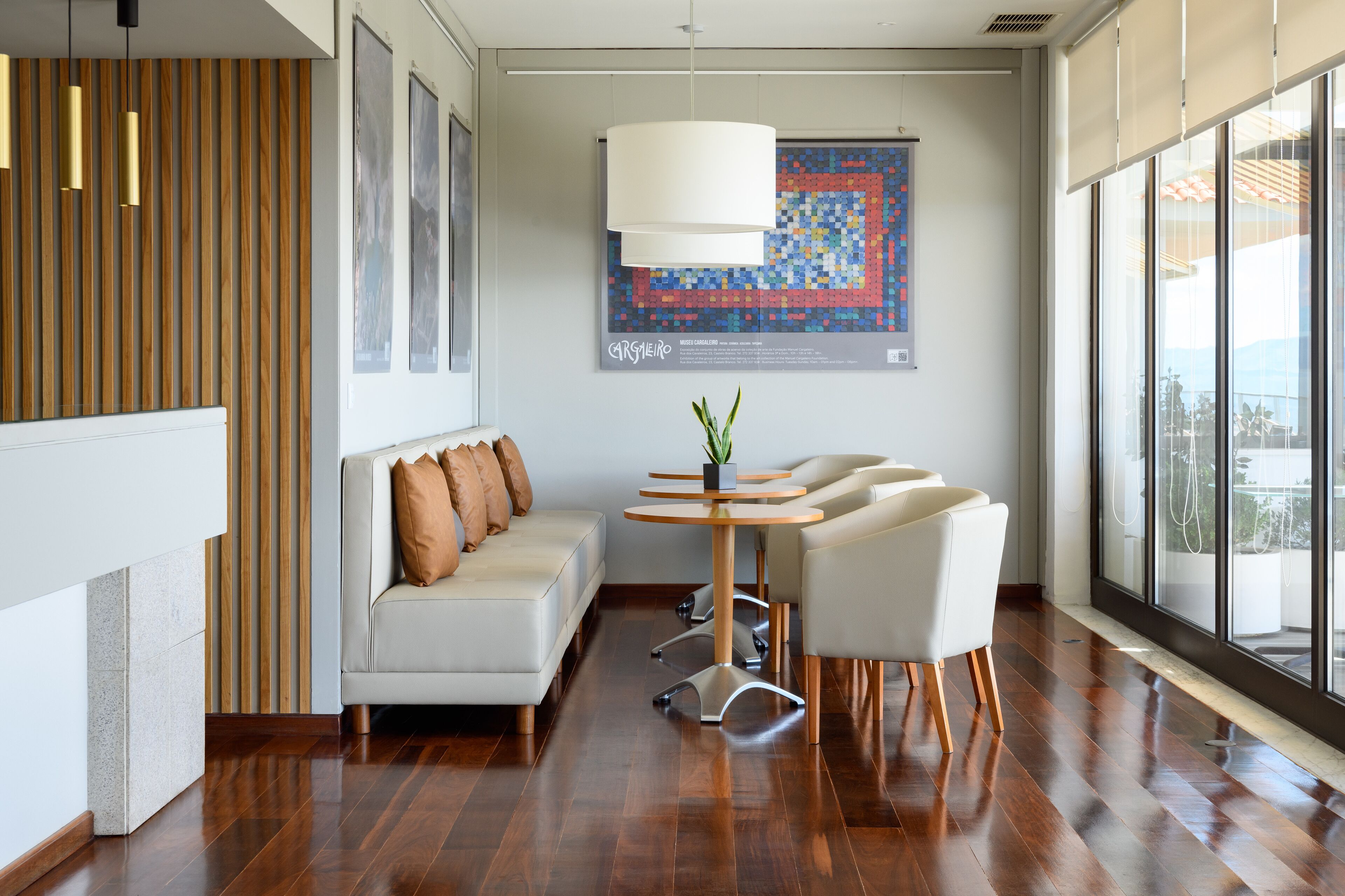 A modern interior with a wooden slat wall, a white sofa with brown cushions, a round table with chairs, and a large window with a view.