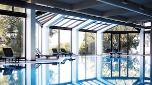 An indoor swimming pool with clear glass walls, sun loungers, and a view of trees outside. - Melia Castelo Branco (Castelo Branco)