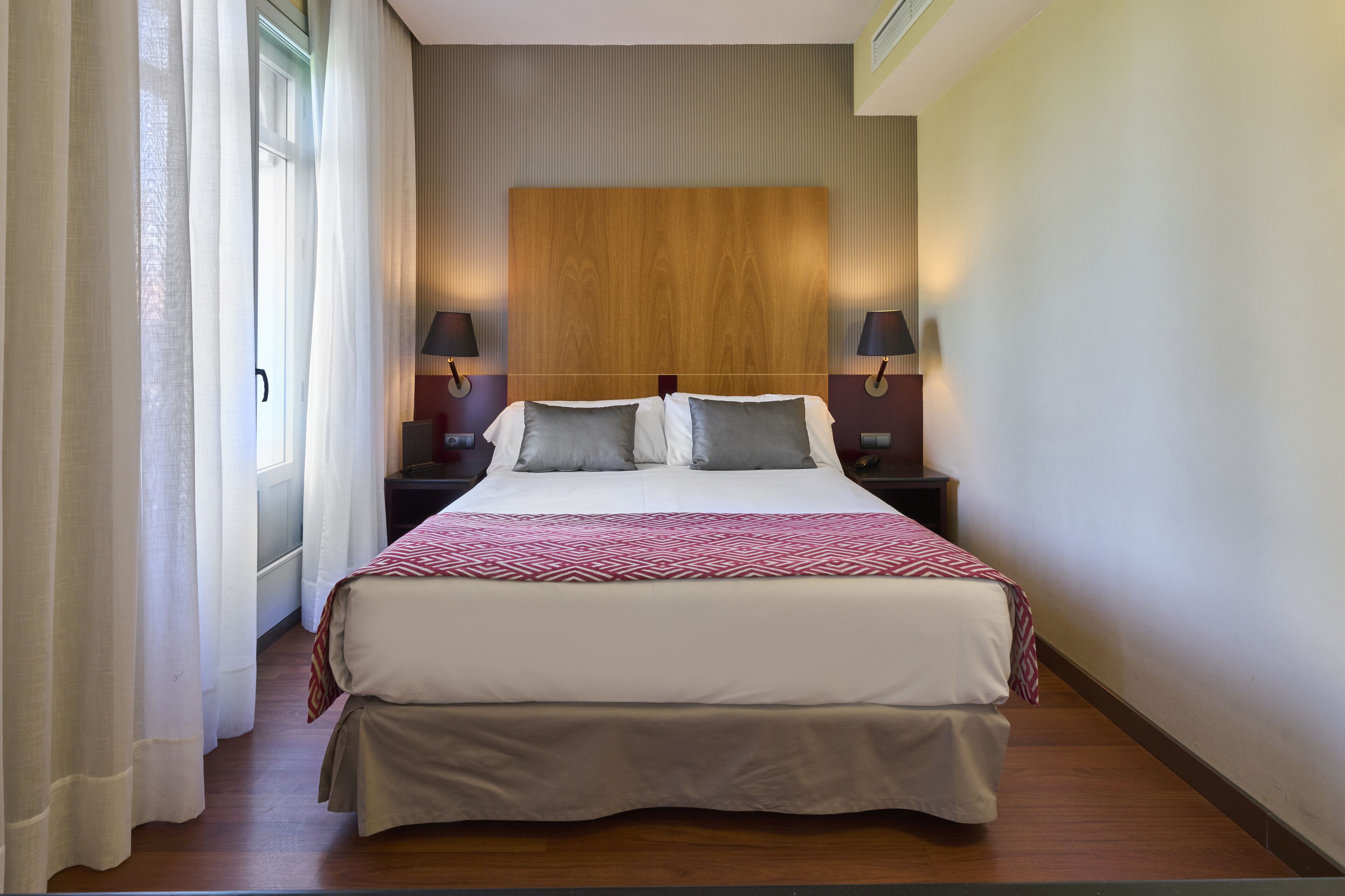 A hotel room with a large bed, two bedside tables with lamps, a wooden headboard, and a window with sheer curtains.
