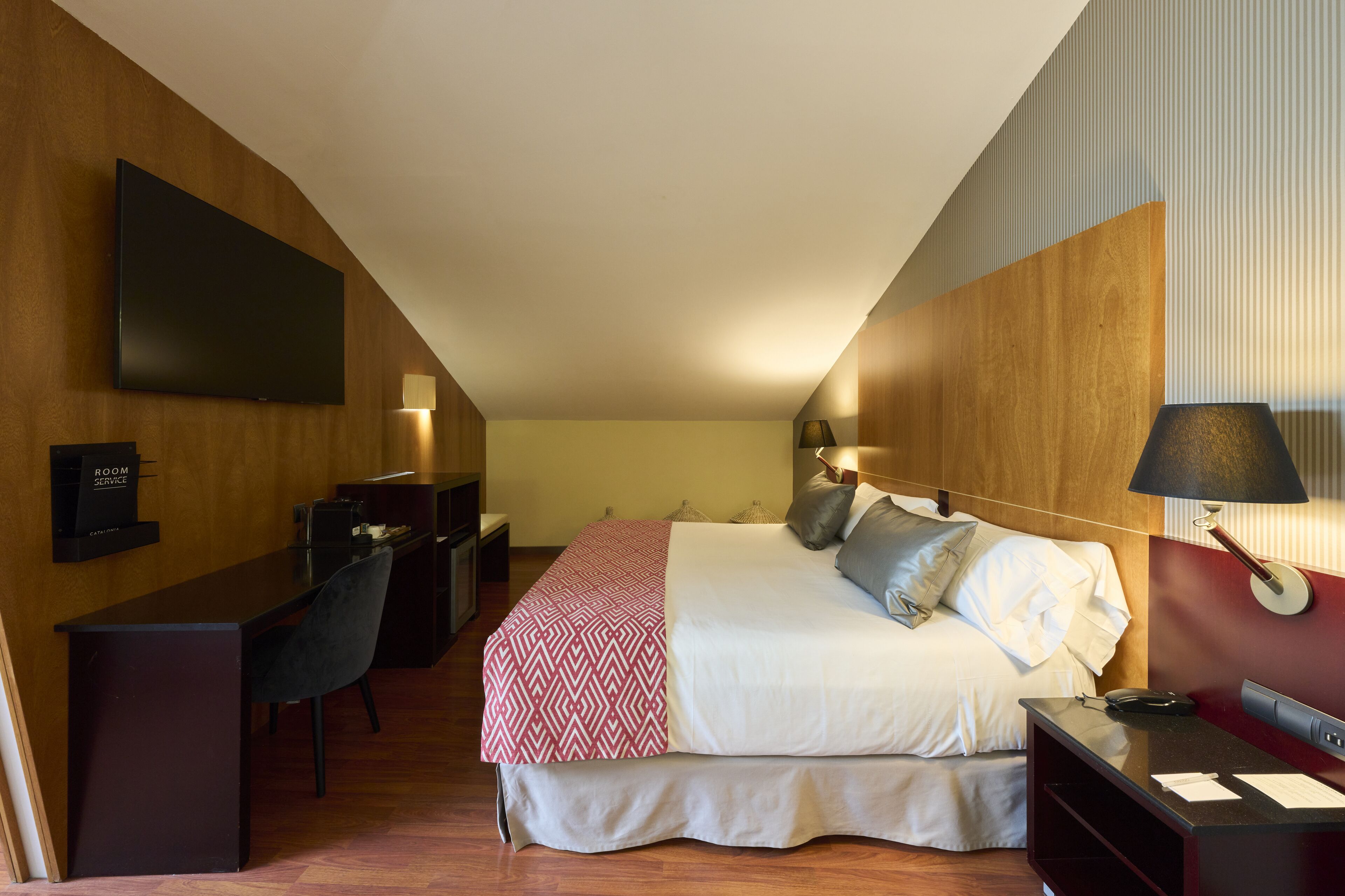 A hotel room with a bed, desk, chair, and a flat-screen TV mounted on the wall.
