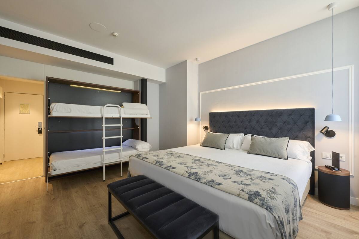 A modern hotel room with a large bed, a bench, a bunk bed, and a bedside table with a lamp.