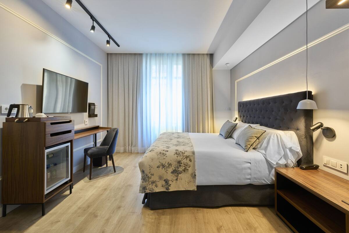 A modern hotel room with a bed, desk, chair, TV, and a large window with curtains.