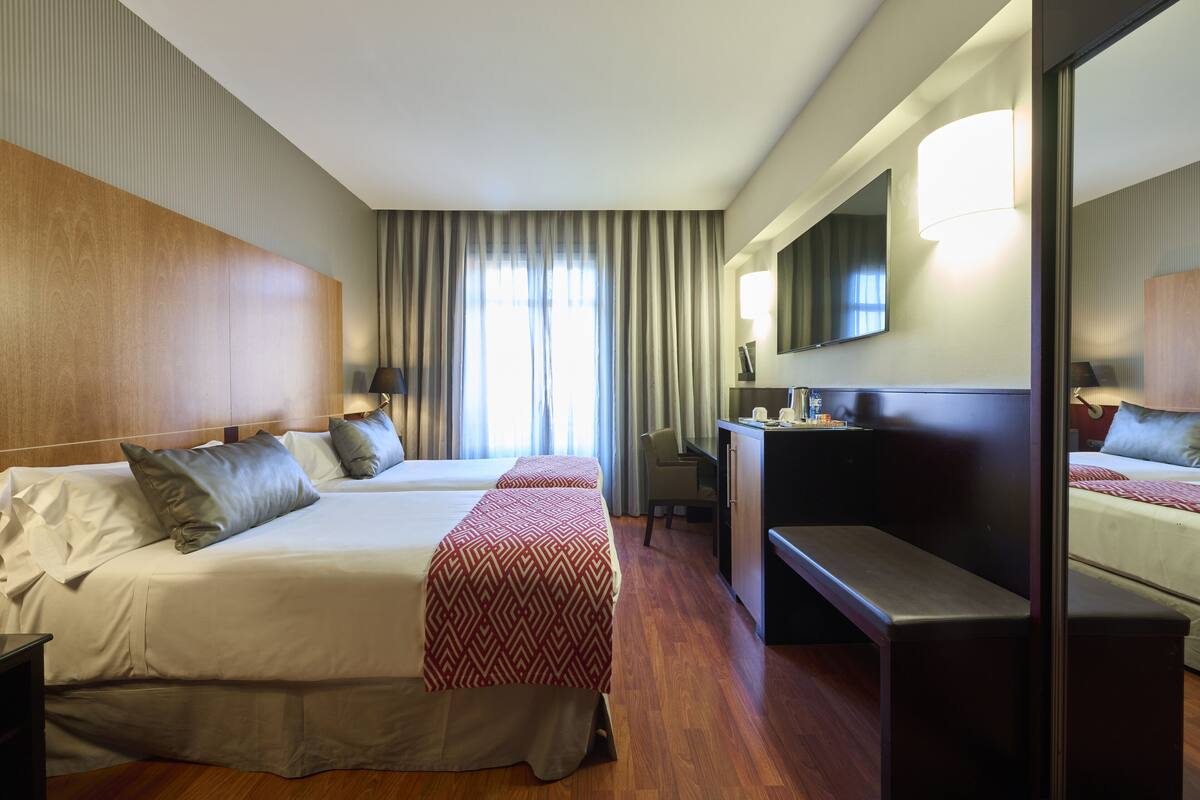 A hotel room with two beds, a desk, a chair, a large mirror, and a window with curtains.