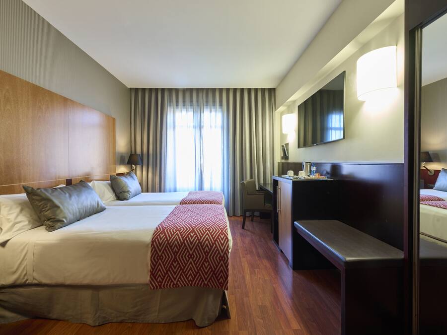 Habitación de hotel con dos camas, un escritorio, una silla, un espejo grande y una ventana con cortinas.