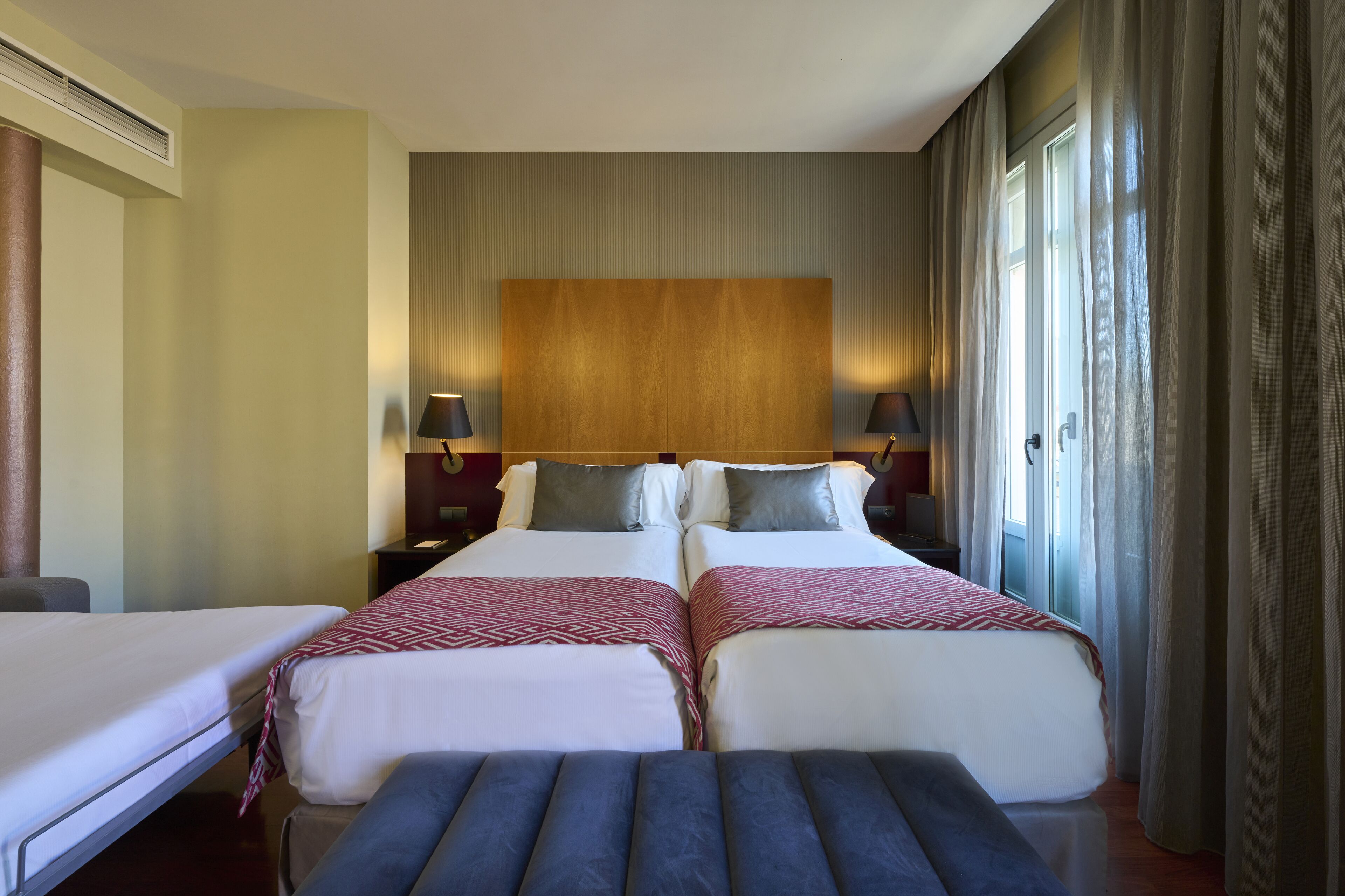 Habitación de hotel con dos camas, un ventanal con cortinas, cabecera de madera y lámparas de noche.