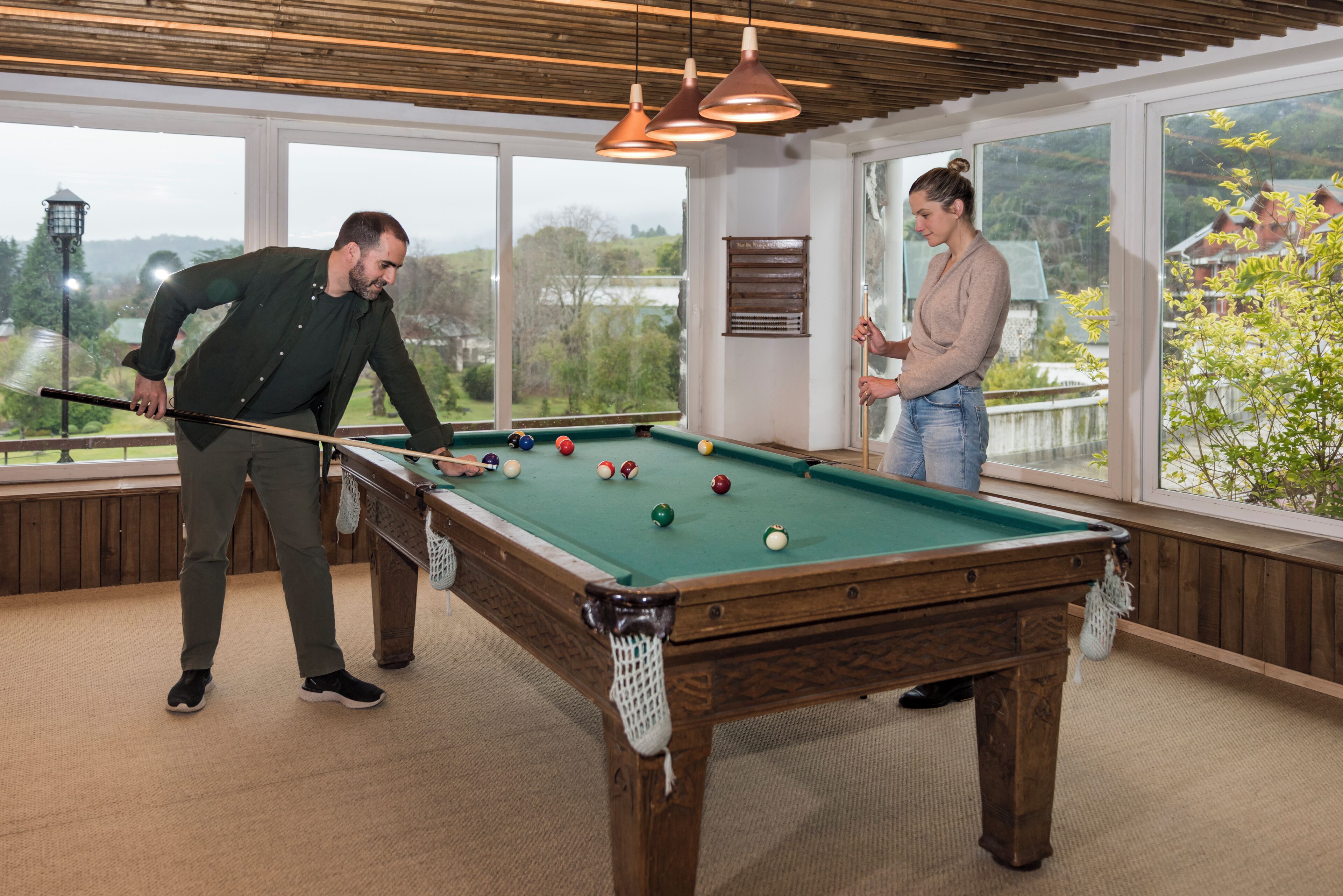 A pool table in a room with large windows, wooden ceiling, and hanging pendant lights.