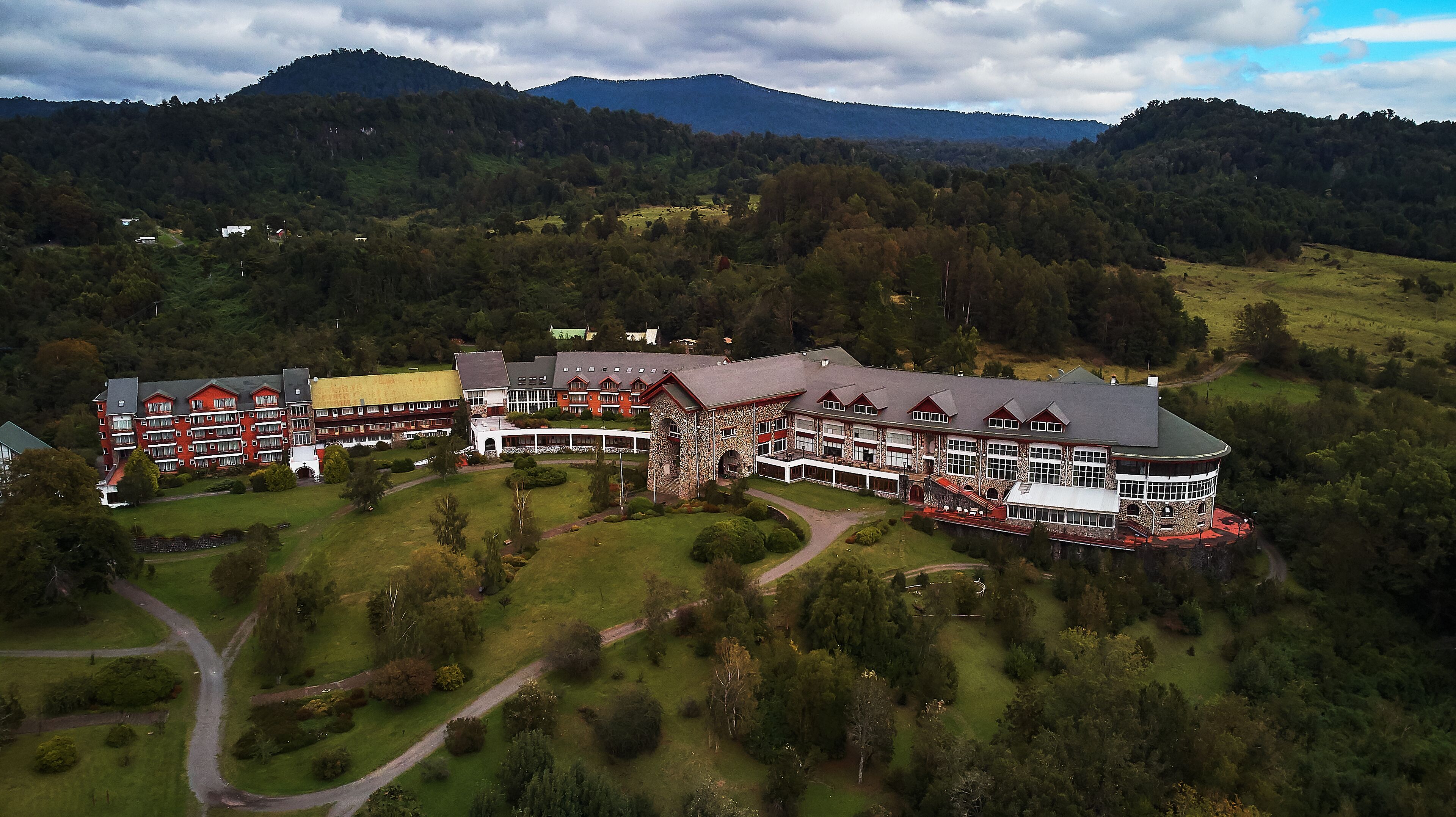 Un hotel grande de varios pisos, con fachada de piedra y múltiples balcones, rodeado de abundante vegetación y colinas.
