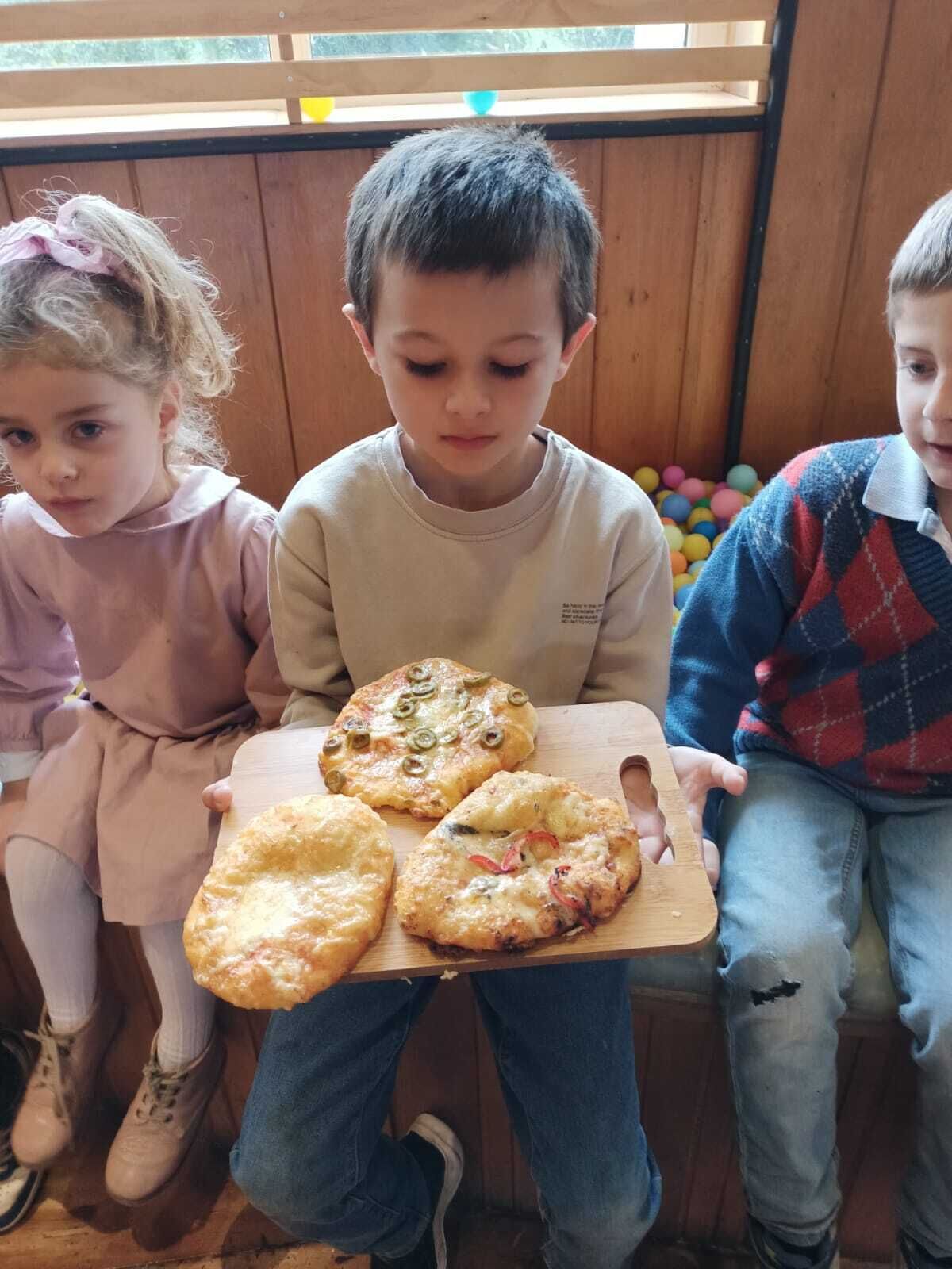 Trois enfants tenant une planche en bois sur laquelle sont posées trois galettes, assis dans une pièce aux murs en bois, avec un espace rempli de balles en arrière-plan.