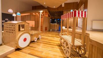 A wooden playhouse with a wagon and a bench, featuring a striped awning and hanging lights.
