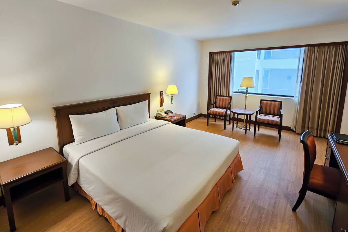 Habitación de hotel con una cama grande, mesitas de noche, un escritorio con silla, una pequeña mesa con dos sillas y una ventana con cortinas.