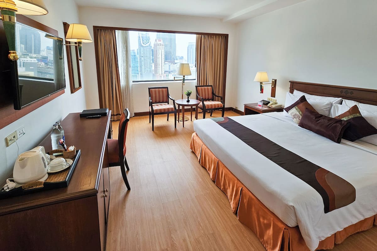A hotel room with a large bed, a desk with a telephone, a chair, a small table with a lamp, and a view of the city through the window.
