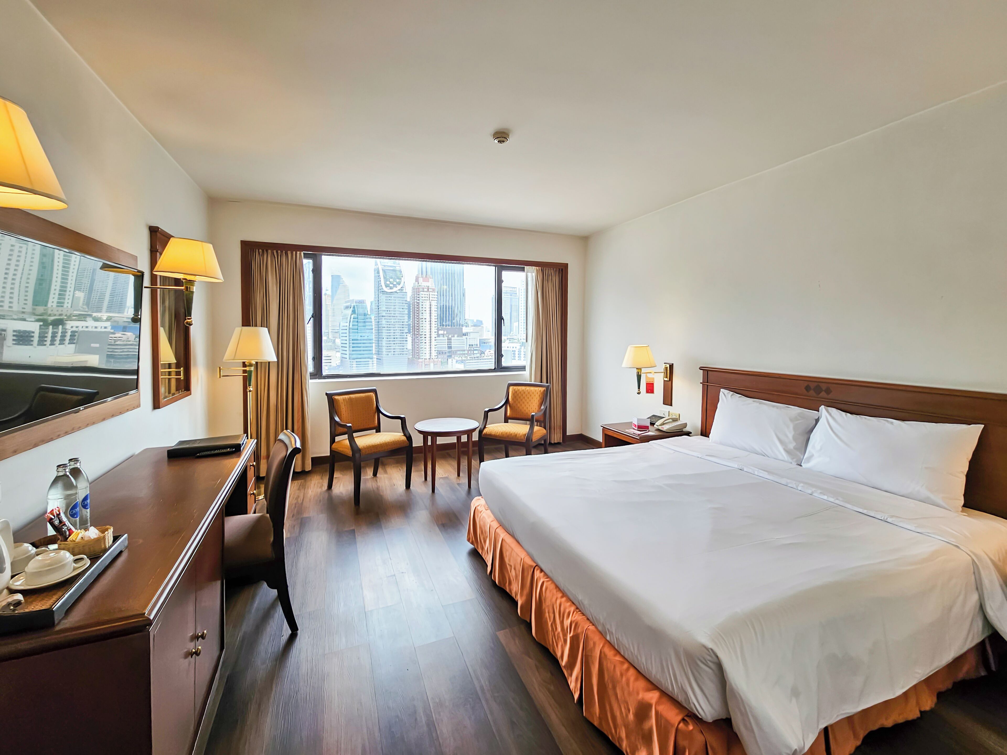 Een hotelkamer met een groot bed, een bureau met stoel, een kleine tafel met twee stoelen, een raam met uitzicht op de stad en een nachtkastje met lamp.