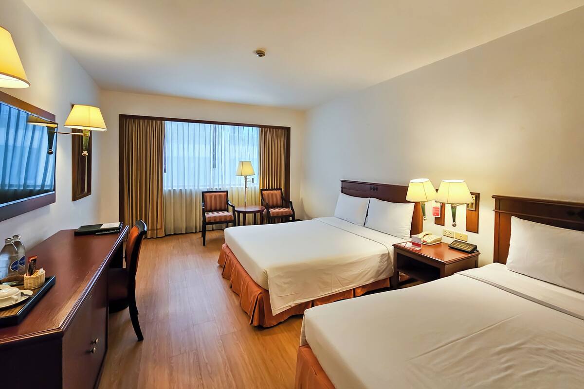 A hotel room with two beds, a desk, a chair, a lamp, and a window with curtains.