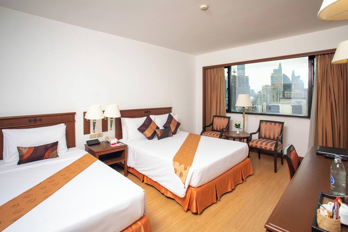 A hotel room with two beds, a desk, a chair, and a large window with a city view.