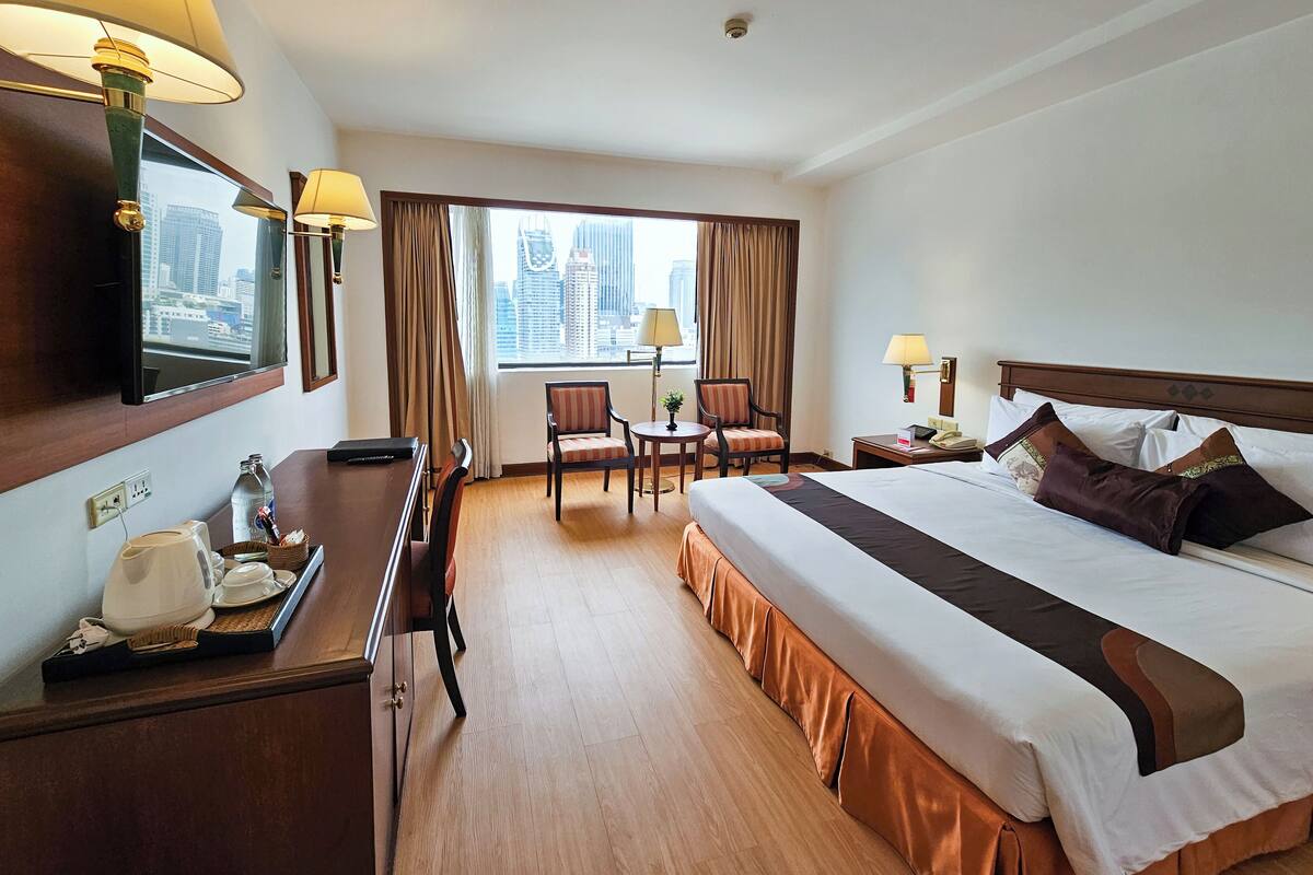 Habitación de hotel con cama, escritorio, silla, mesita, lámpara y ventana con vistas a edificios.