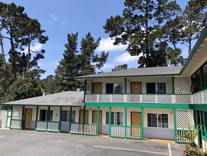 A two-story motel with a green and white color scheme, featuring a covered parking area and a view of surrounding trees.