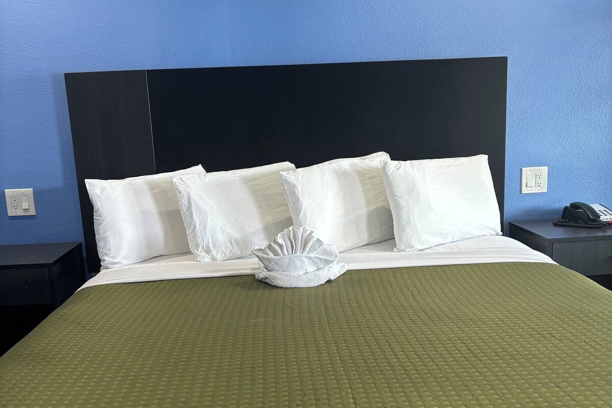 A neatly made bed with white linens and a green bedspread, a black headboard, and a white wall with a power outlet.